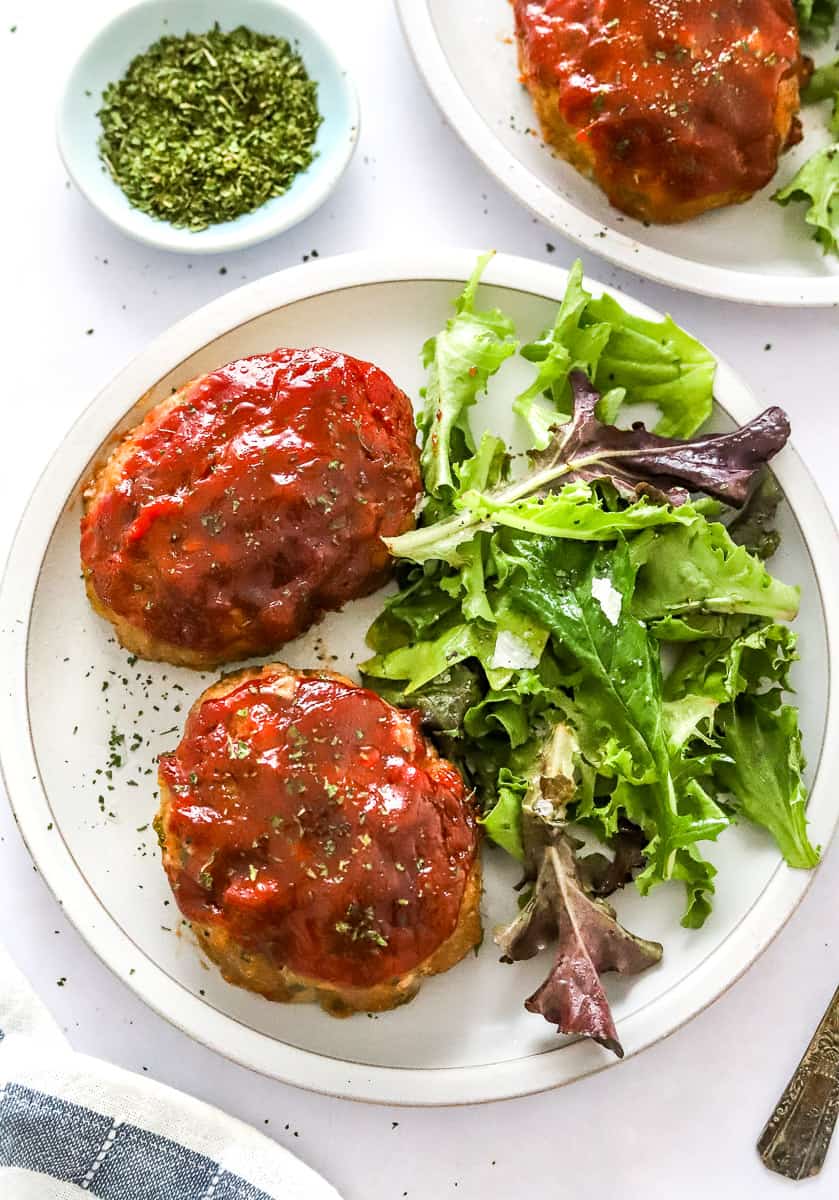 Plate filled with cooked individual meatloafs with mixed greens next to it with another plate of it and bowl of herbs behind it.