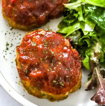 Close up of a small meatloaf topped with a ketchup glaze with greens on the plate next to it.