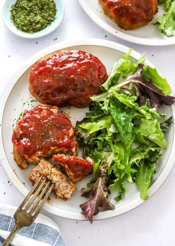 Plate with turkey meatloaves and mixed greens on it with a fork cutting into one of the loaves and another plate of it behind it.