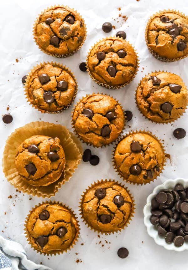 A bunch of chocolate chip muffins on white parchment paper with a white dish with chocolate chips in it in front of it.