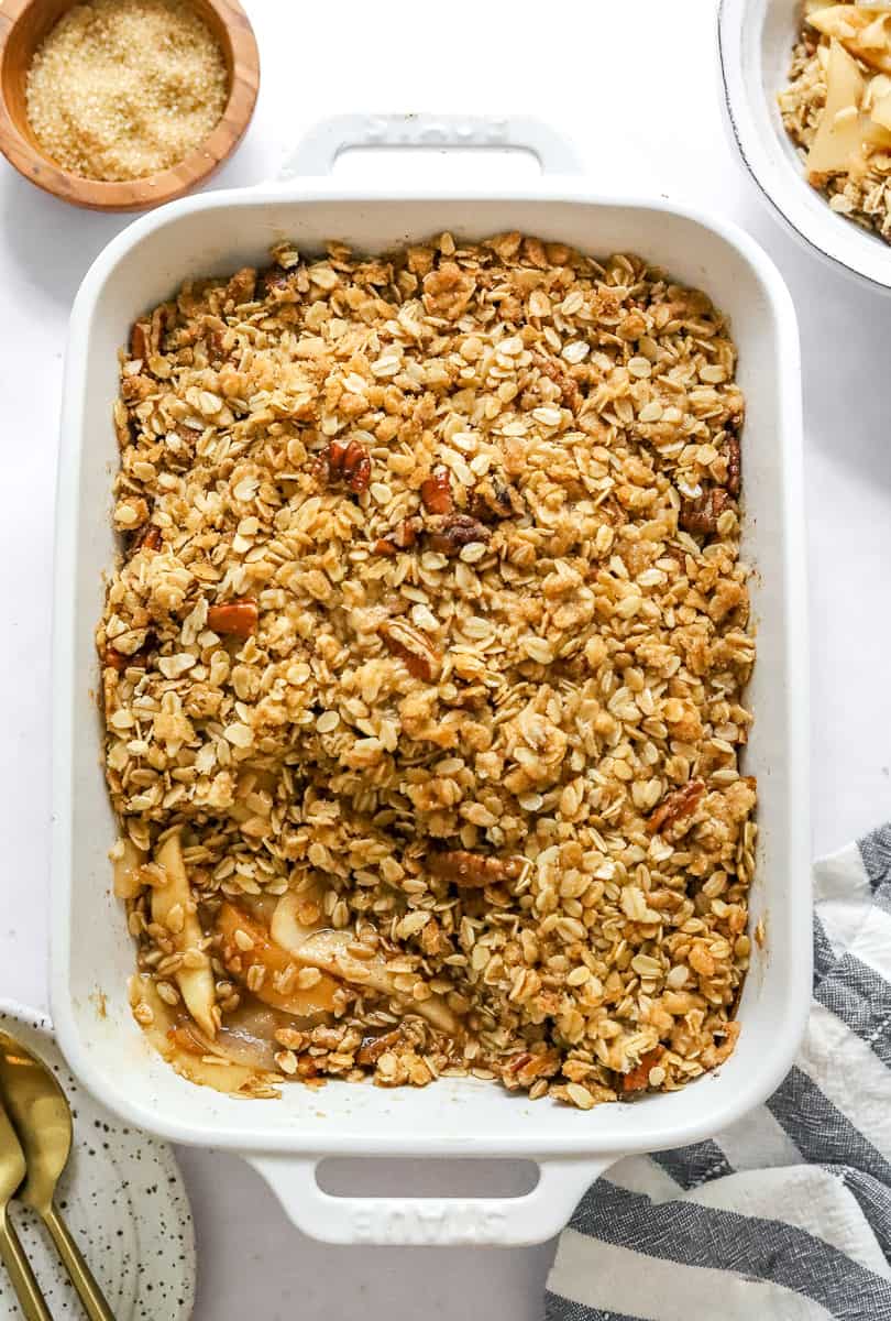 Rectangle white baking pan filled with cooked apples and pears topped with an oat and nut crumble topping with a striped linen in front of it and a bow of sugar and more crisp behind it.