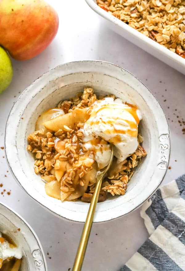 Bowl filled with apple pear crisp topped with vanilla ice cream with a gold spoon in the bowl and crisp and apples behind it.