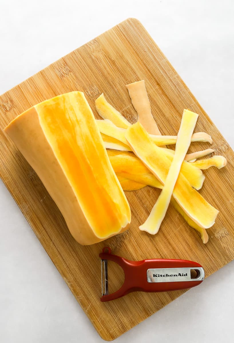 Cutting board with the neck of a butternut squash on it half peeled with a red vegetable peeler on the board in front of it. 