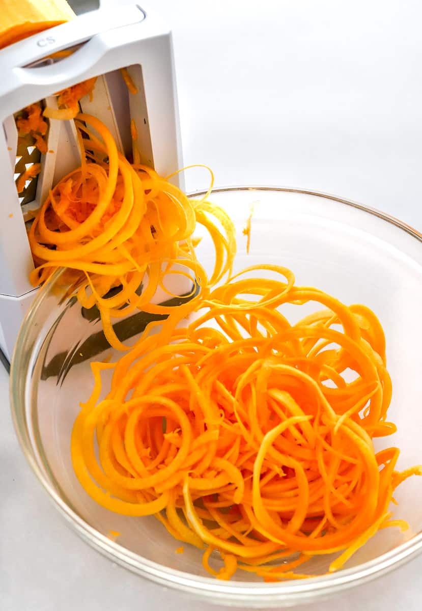 Spiralizer with butternut squash noodles in it with a bowl of butternut squash noodles in front of it. 