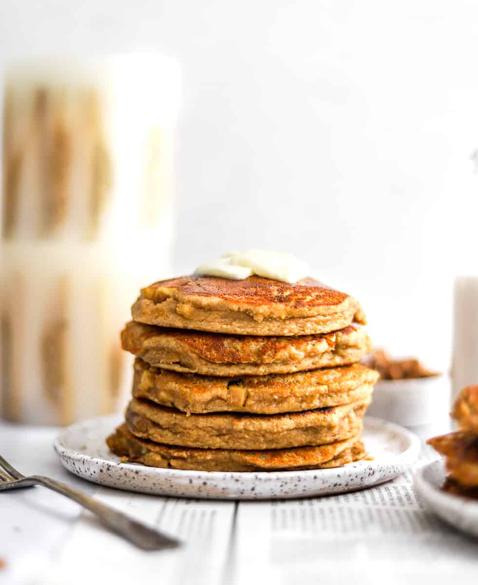 Apple cinnamon pancakes on a plate with a fork and more pancakes in front of them and a candle and bowl of nuts behind them.