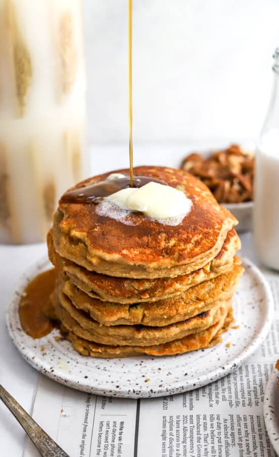 Stack of cinnamon pancakes with butter on them with syrup being pour on them on a plate on top of newspaper with candles and a bowl of nuts behind it.