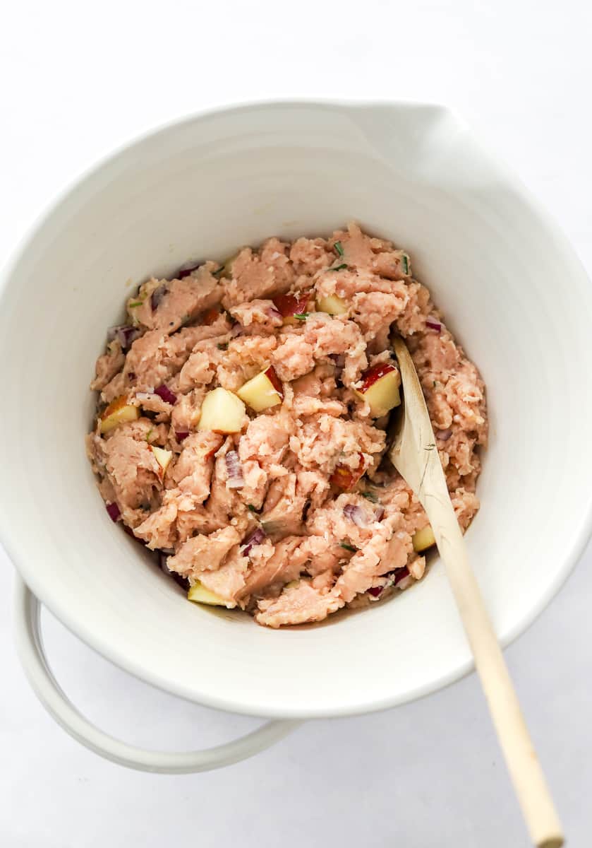 Mixing bowl filled with uncooked ground chicken mixed with apple chunks in it with a wooden spoon in the bowl.
