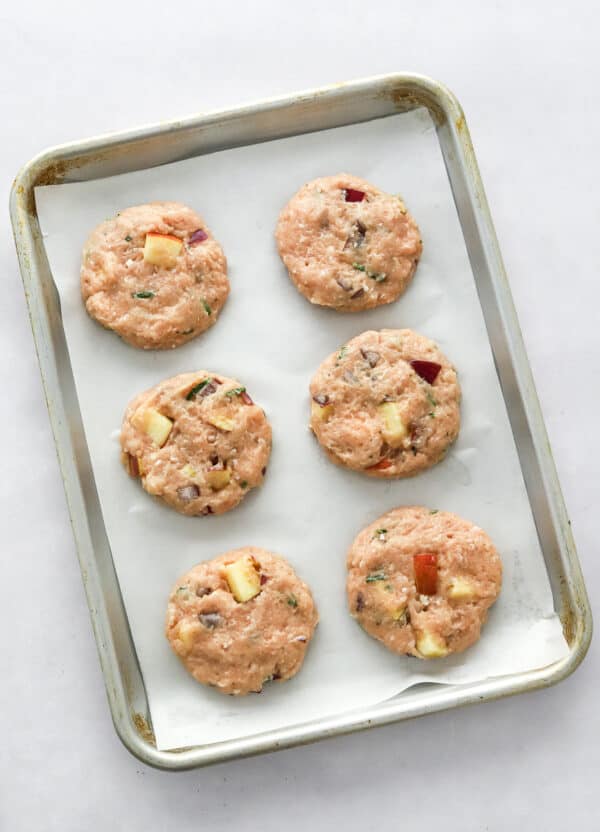 Formed ground chicken sausages with apple and onion in them on a baking sheet.