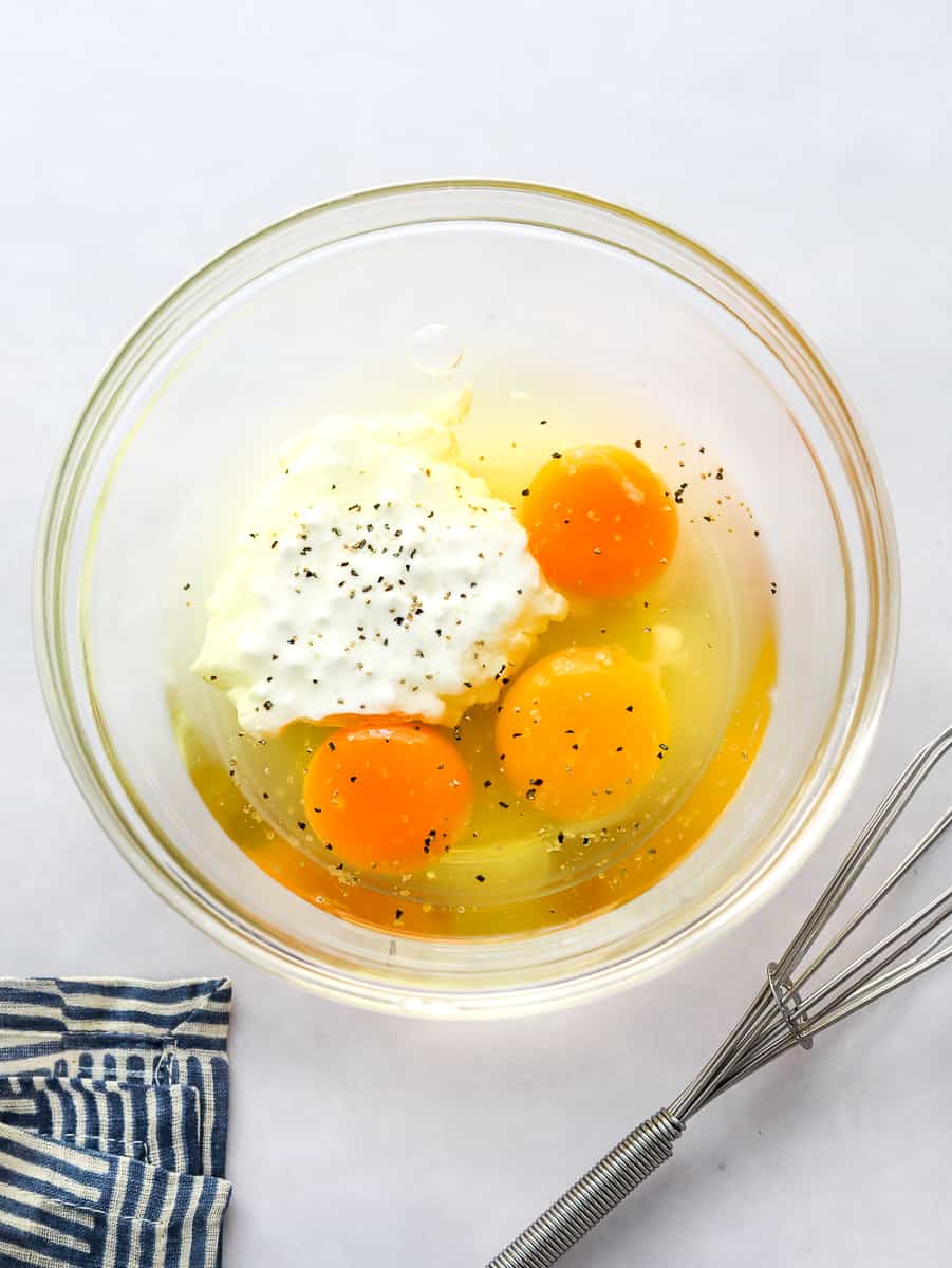Glass mixing bowl with cracked eggs and some cottage cheese in it with a whisk next to the bowl and a stripped towel in front of it.