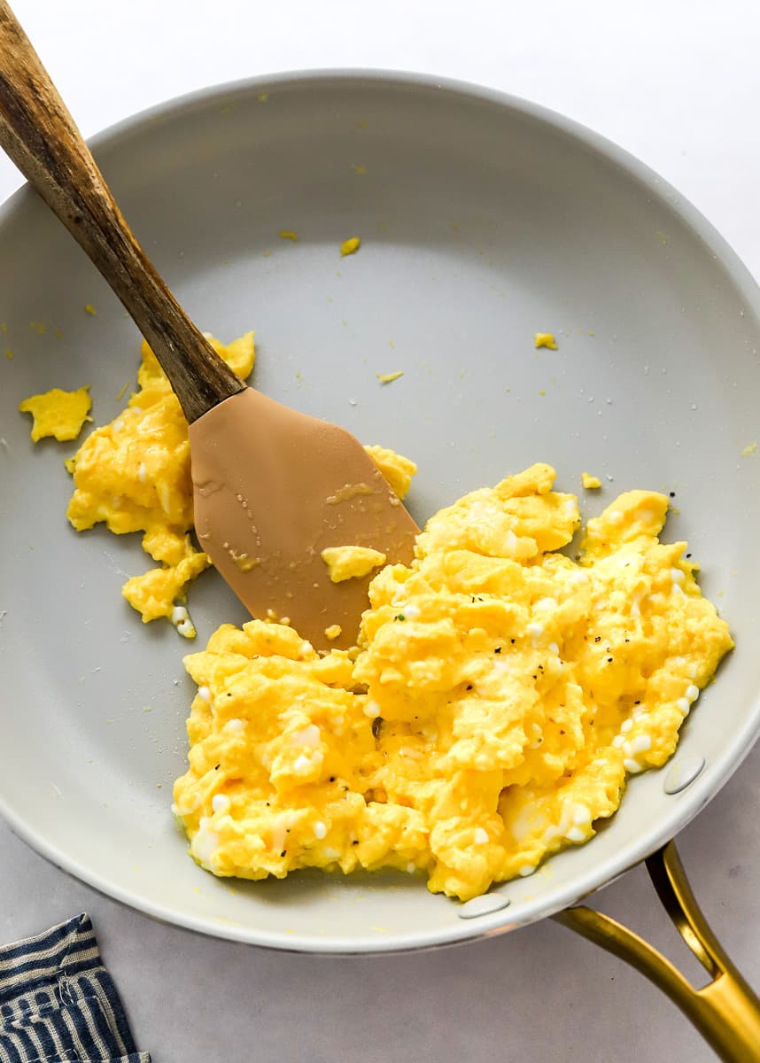 Cooked scrambled cottage cheese eggs in a grey pan with a rubber spatula in the pan with a stripped towel in front of it.