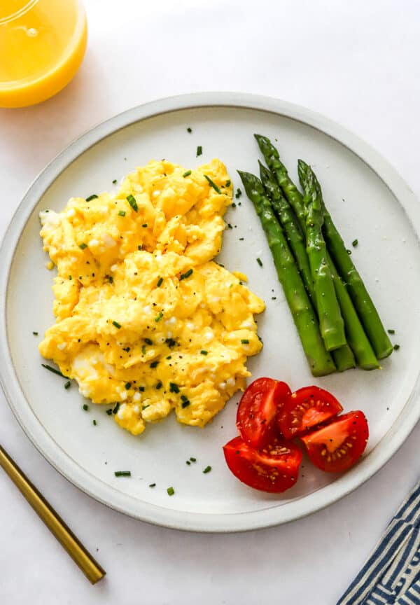 Cottage cheese eggs on a plate with asparagus and chopped tomatoes with a stripped towel and gold fork in front of it and a glass of orange juice behind it.