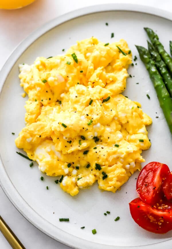 Scrambled eggs topped with chopped chives on a plate with asparagus and chopped tomatoes.