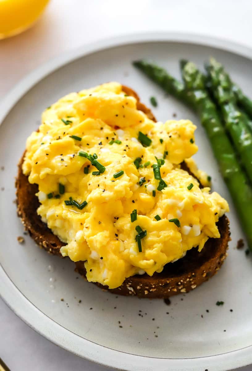 Scrambled eggs on toast on a plate with asparagus next to it on the plate and a glass of juice behind it.