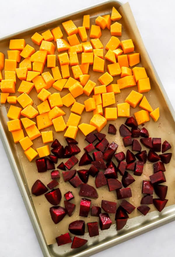 Baking sheet lined with brown parchment paper with uncooked cubes of squash and beets on the pan.