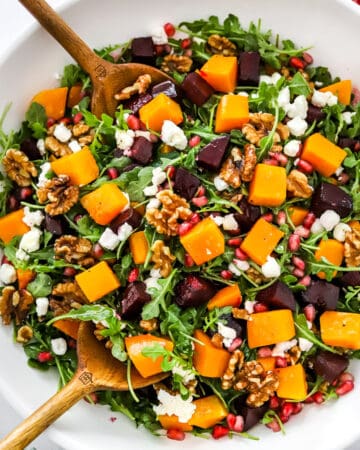Round, white salad bowl filled with roasted butternut squash salad with beets, nuts and cheese with wood salad serving spoons in the bowl and a green linen in front of it.