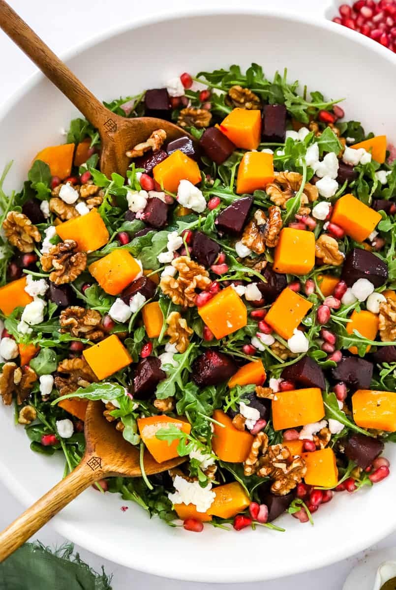 Round, white salad bowl filled with roasted butternut squash salad with beets, nuts and cheese with wood salad serving spoons in the bowl and a green linen in front of it.