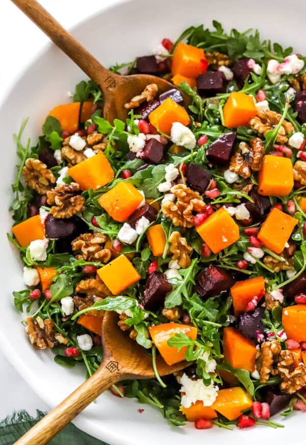 Salad bowl filled with arugula, and cubes of roasted beets and squash with serving spoons in the bowl.
