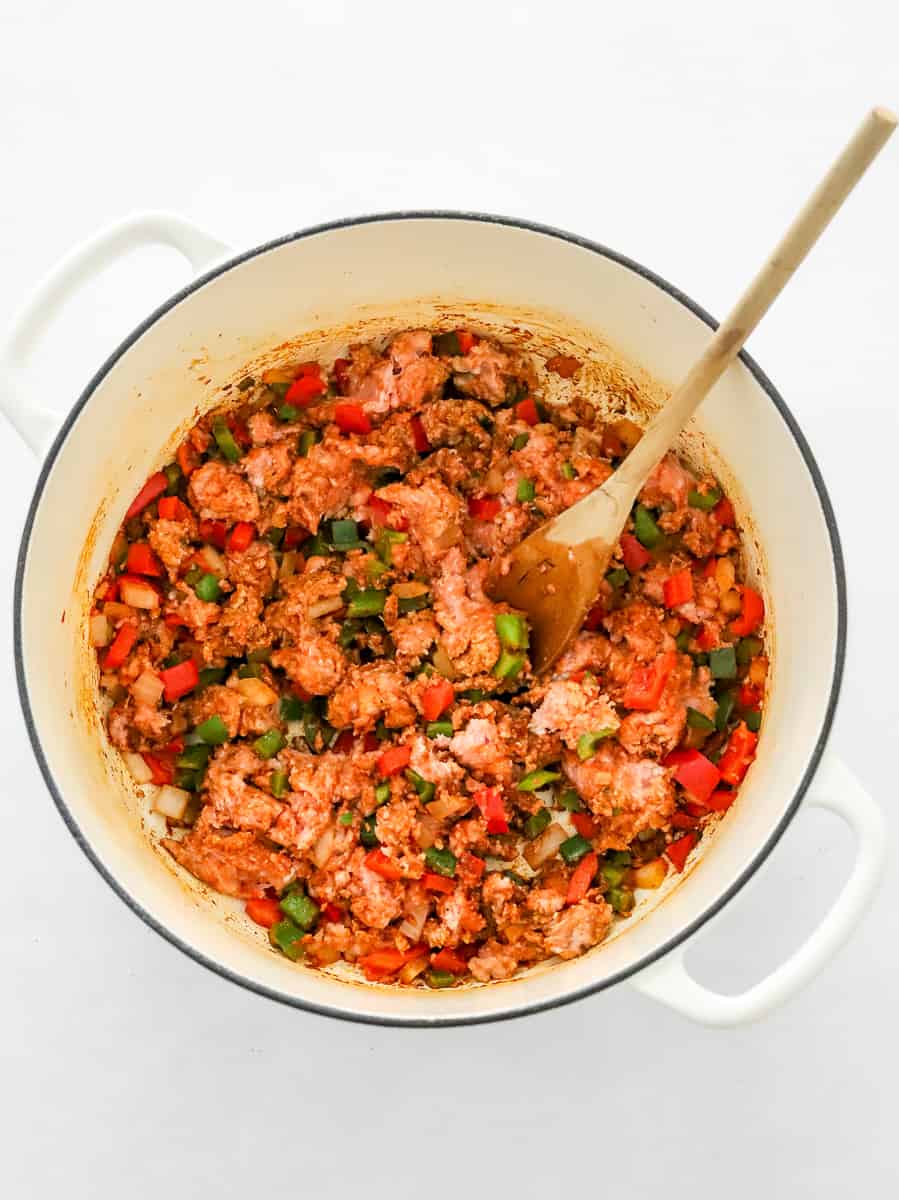 White pot with raw ground turkey and diced peppers and onions in it with a wooden spoon in the pot. 