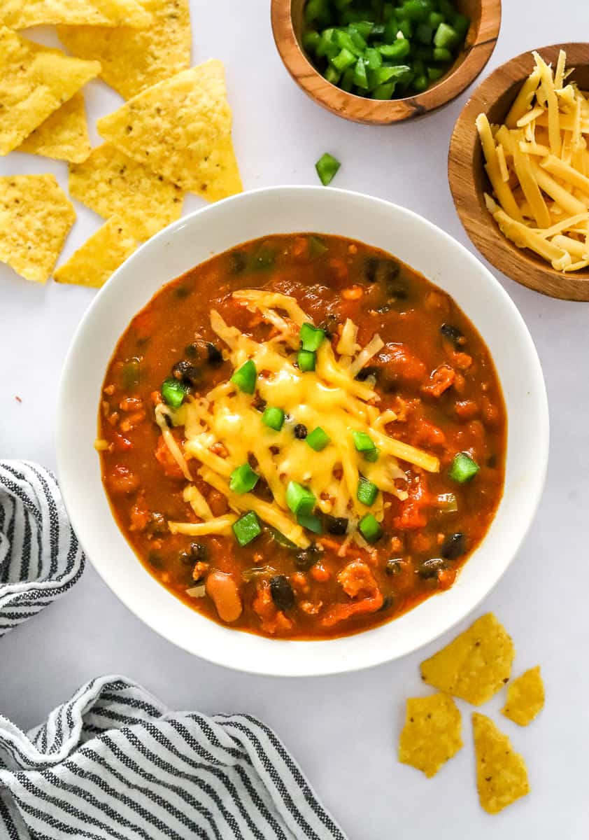 Pumpkin chili with turkey in a bowl topped with melted cheese and green peppers with tortilla chips and a bowl of shredded cheese and peppers behind it. 