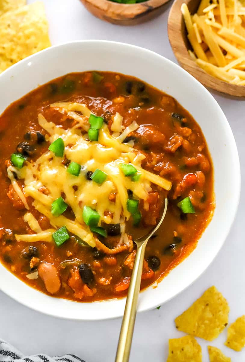 Pumpkin chili in a white bowl with melted cheese on it and diced pepper with a spoon in the bowl and tortilla chips around it. 