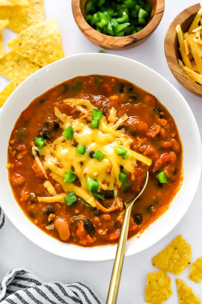 Chili in a white bowl with melted cheddar cheese ontop and a spoon in the bowl with tortilla chips and more cheese and chopped green peppers behind it. 