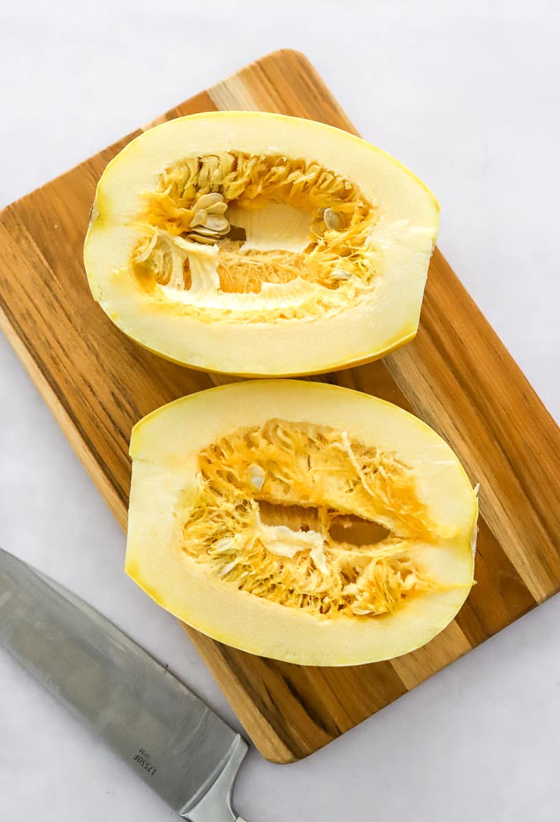 Uncooked spaghetti squash cut in half on a wood cutting board with a silver knife next to it.