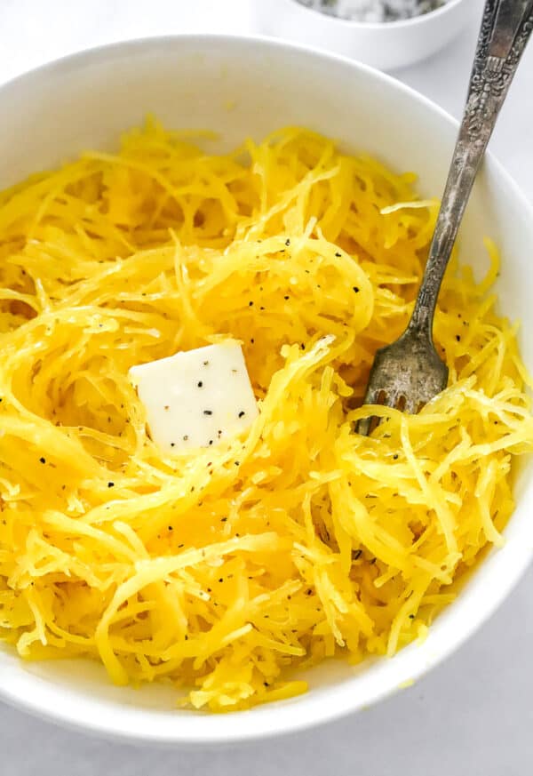 Cooked spaghetti squash in a white serving bowl with butter on it wit ha metal fork in the bowl with the squash.