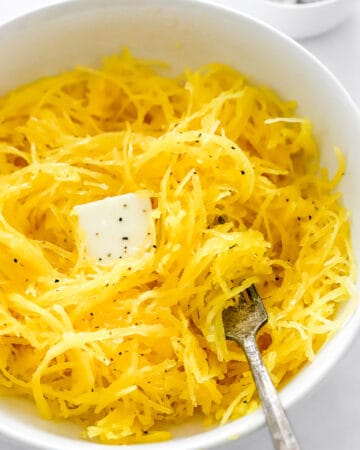 Microwave spaghetti squash in a bowl with butter and salt and it with a fork in the bowl with it and salt and pepper in a bowl behind it.