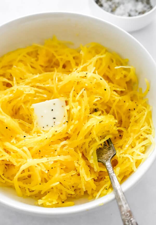Microwave spaghetti squash in a bowl with butter and salt and it with a fork in the bowl with it and salt and pepper in a bowl behind it.