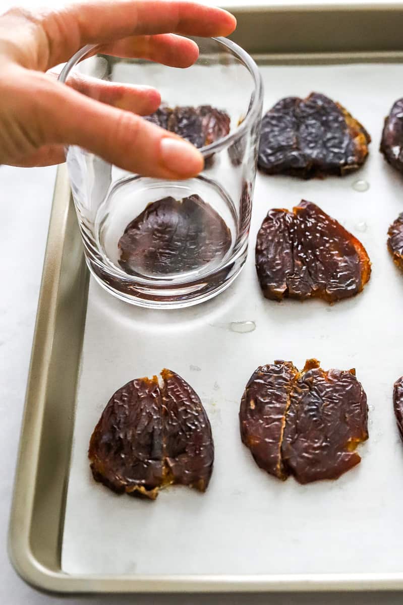 Dates on a lined baking sheet with a hand using a glass to prep one of them flat.