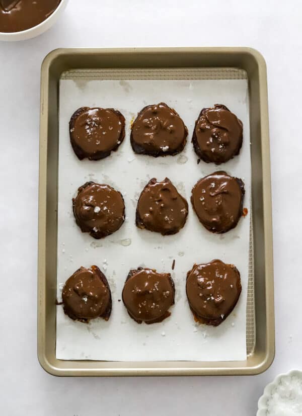Lined baking sheet with date cookies topped with melted chocolate on it with a bowl of more melted chocolate behind it.