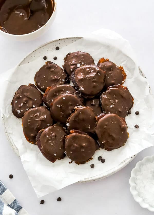 A bunch of date chocolate cookies on a plate with a bowl of sea salt in front of it and more melted chocolate in a bowl behind it.