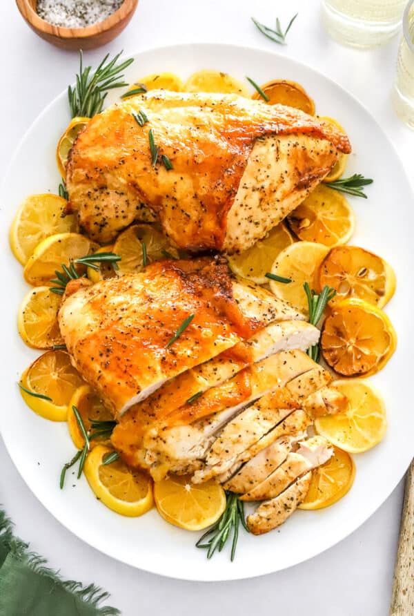 Two golden cooked turkey breast on a white platter with sliced lemons and herbs around the turkey.