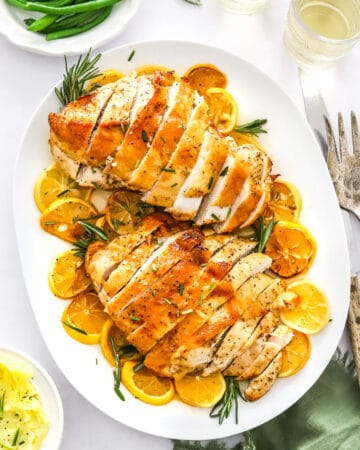 Platter of two boneless turkey breast sliced on top of lemon slices with some rosemary with mashed potatoes on a plate in front of it and a bowl of green beans and glasses of white wine behind it.