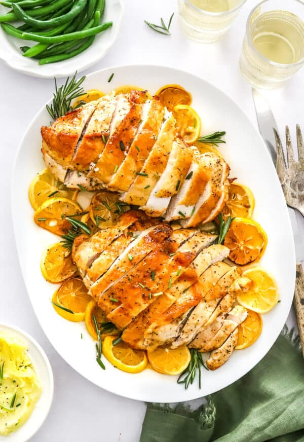 Platter of two boneless turkey breast sliced on top of lemon slices with some rosemary with mashed potatoes on a plate in front of it and a bowl of green beans and glasses of white wine behind it.