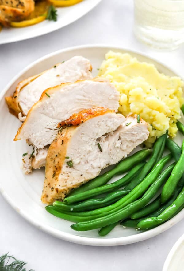 Sliced roasted turkey on a plate with green beans and mashed potatoes with more turkey and a glass of wine behind it.