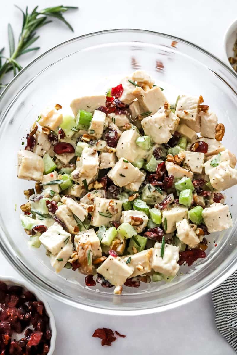 Mixed up cranberry pecan turkey salad in a glass bowl with rosemary and more nuts behind it. 