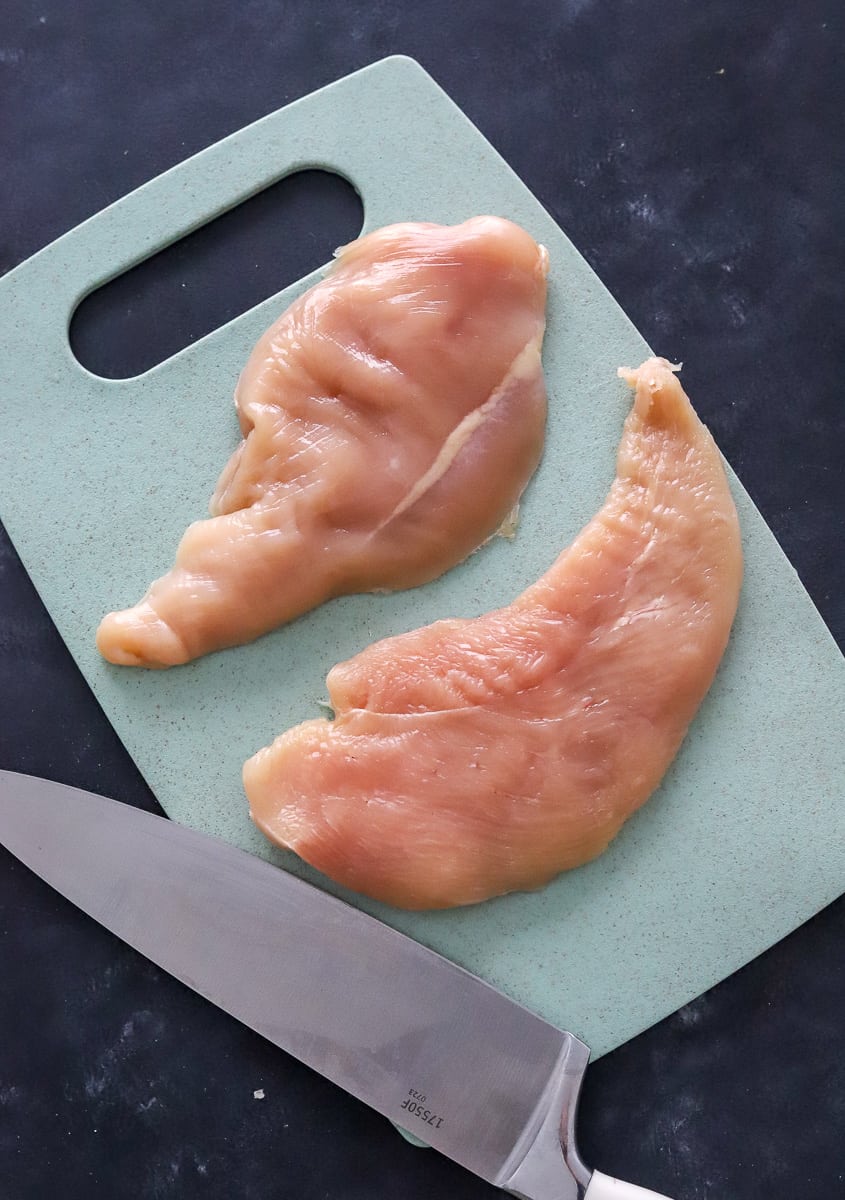 Raw boneless chicken breast halves on a blue cutting board with a white and silver kitchen knife in front of it. 