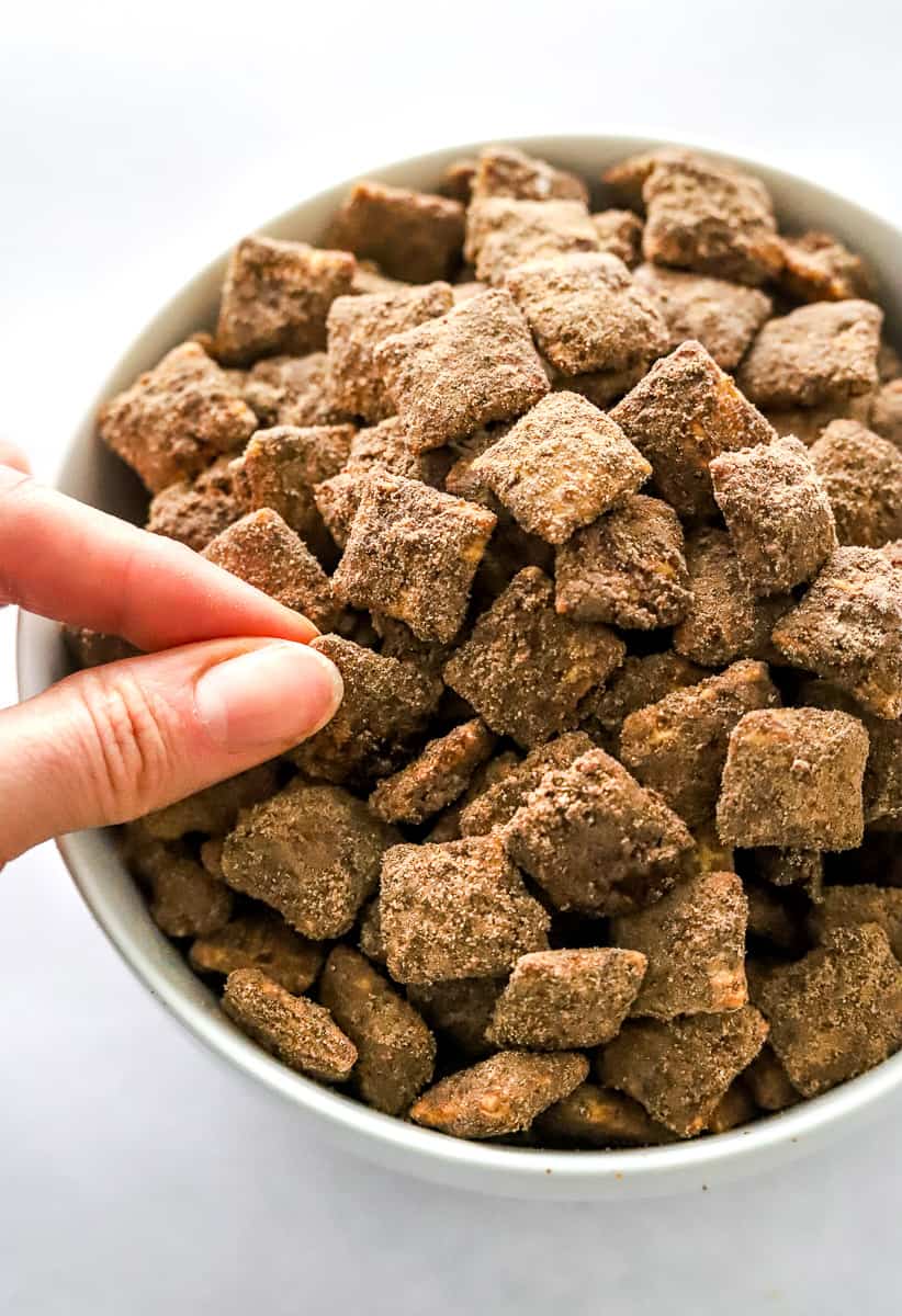 Hand grabbing a piece of chocolate peanut butter covered Chex mix in a white bowl.
