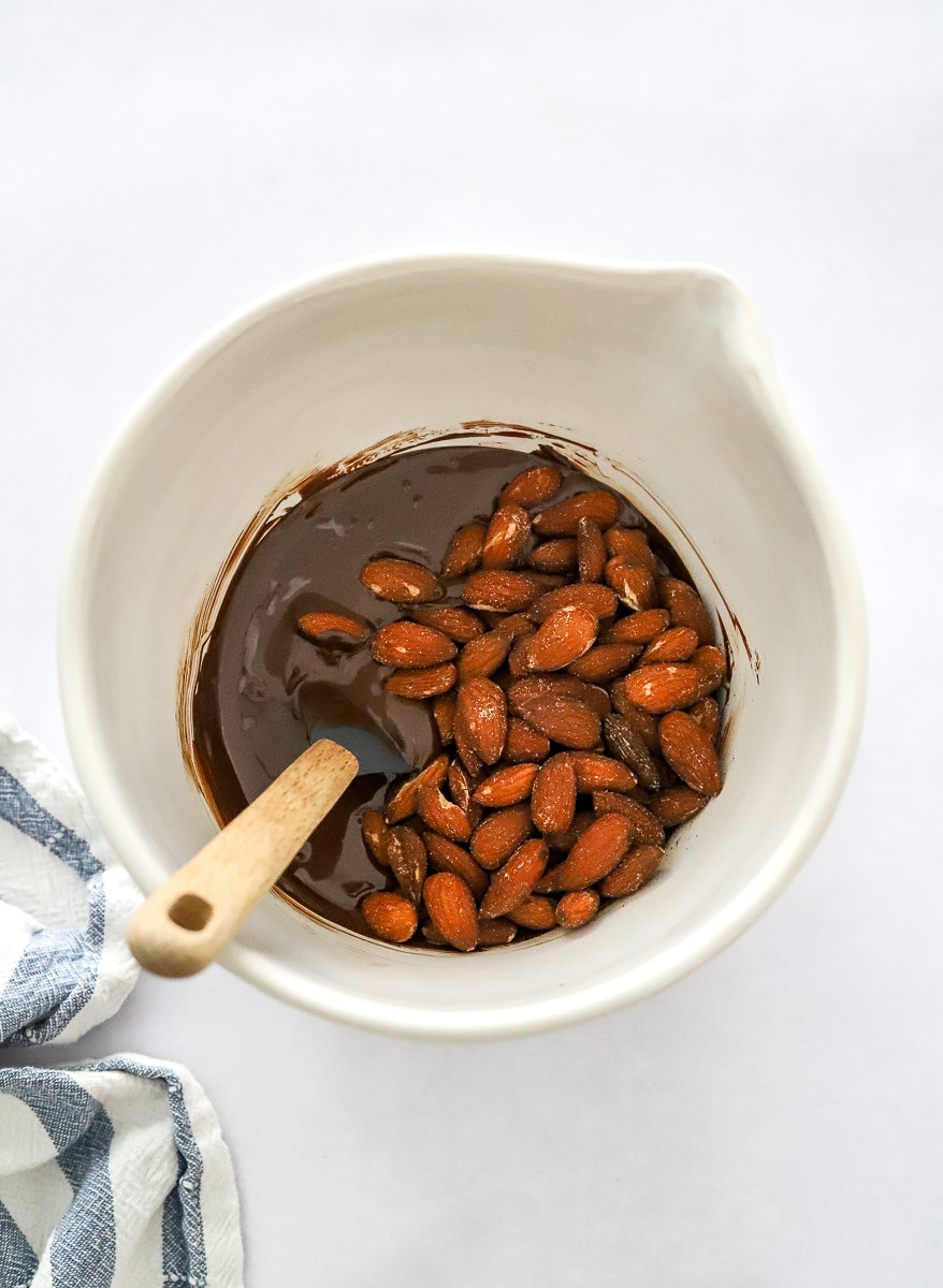 Melted dark chocolate with almonds in a mixing bowl with a small rubber spatula in it and a striped towel in front of it. 