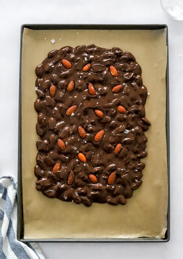 Lined baking sheet with dark chocolate mixed with almonds spread like bark on it before setting with a striped blue and white towel in front of it.