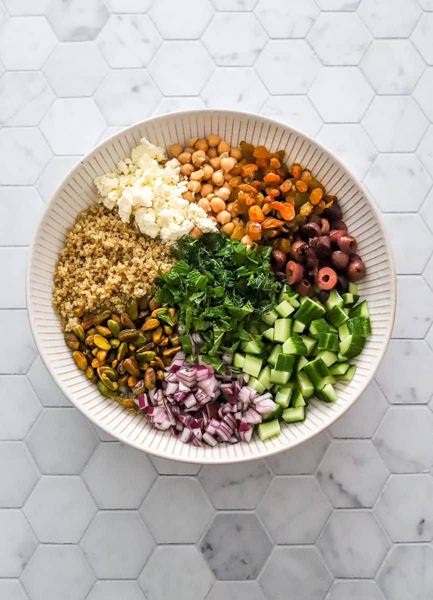 Quinoa, olives, onion, cucumber, nuts, feta, herbs and chickpeas in a white bowl.