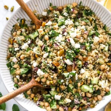 Viral Jennifer Aniston salad with chickpeas, veggies, nuts feta and a lemon dressing with wood serving spoons in the bowl and lemon sliced behind it.