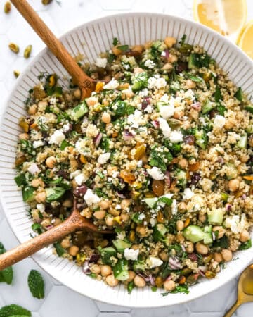 Viral Jennifer Aniston salad with chickpeas, veggies, nuts feta and a lemon dressing with wood serving spoons in the bowl and lemon sliced behind it.