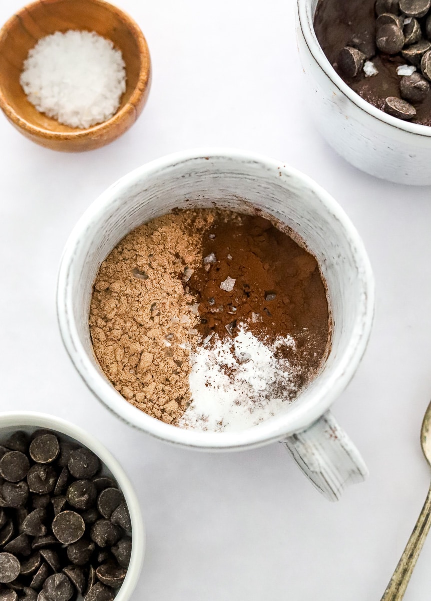Chocolate protein powder, cocoa powder, and baking powder in a mug with a bowl of chocolate chips in front of it and a bowl of salt and another mug of cake behind it.