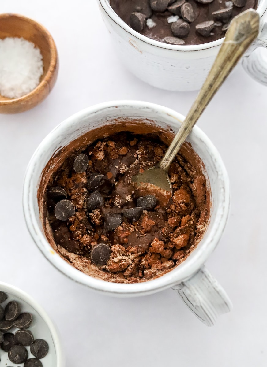 Blended chocolate protein powder with wet ingredients and chocolate chips and a spoon in a mug with another one and salt in a brown dish behind it.