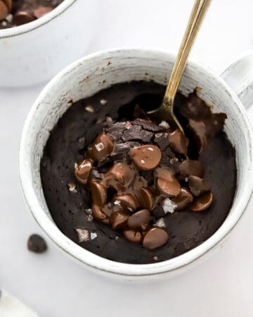 Chocolate cake cooked in a mug with melted chocolate chips and sea salt on top With a spoon in the cake.