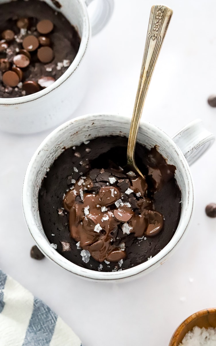 Chocolate protein cake in a mug topped with melted chocolate and salt with a spoon in the mug and another mug cake behind it. 