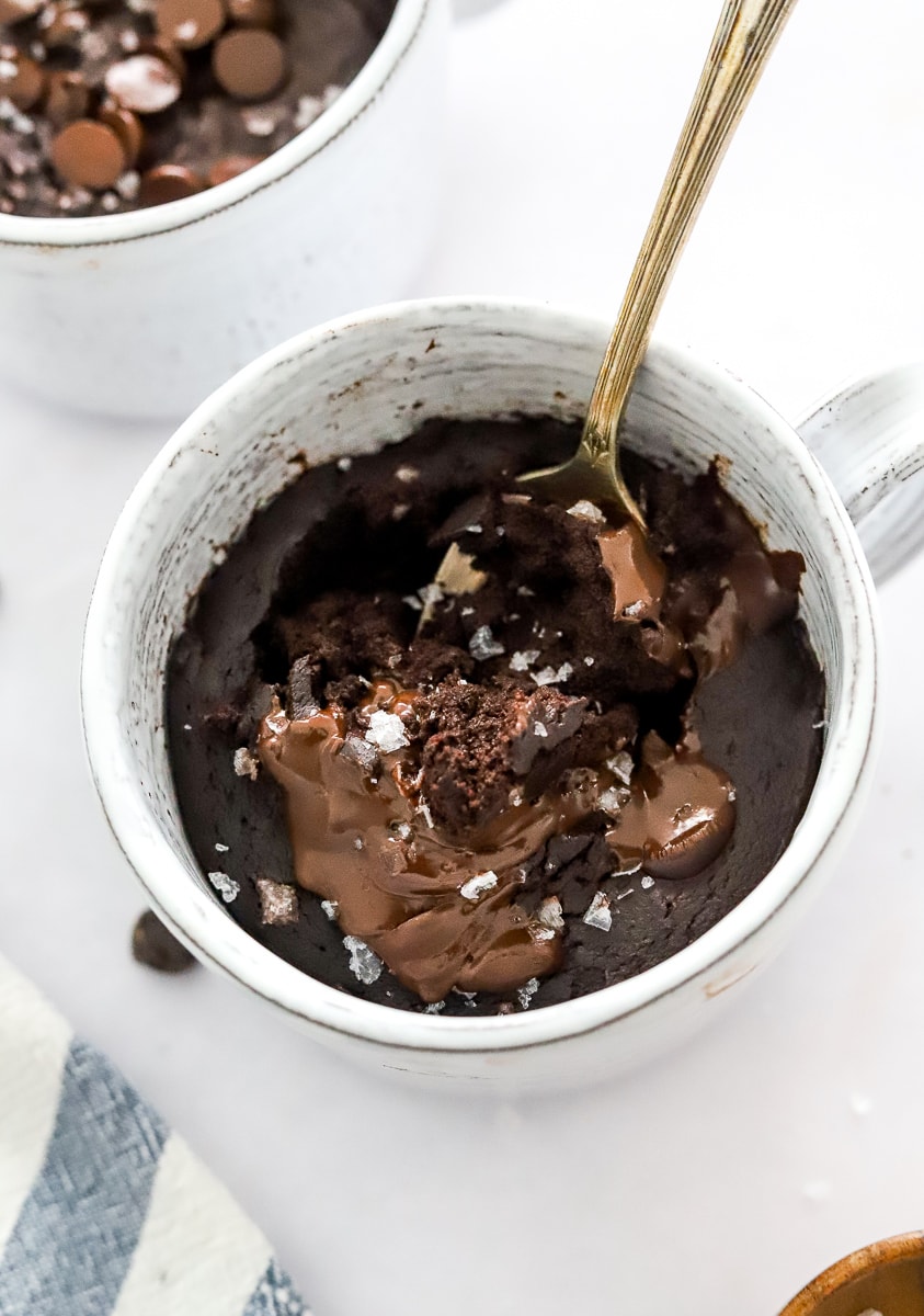 Chocolate cake with melted chocolate on top in a mug with a spoon in cake and another one behind it.
