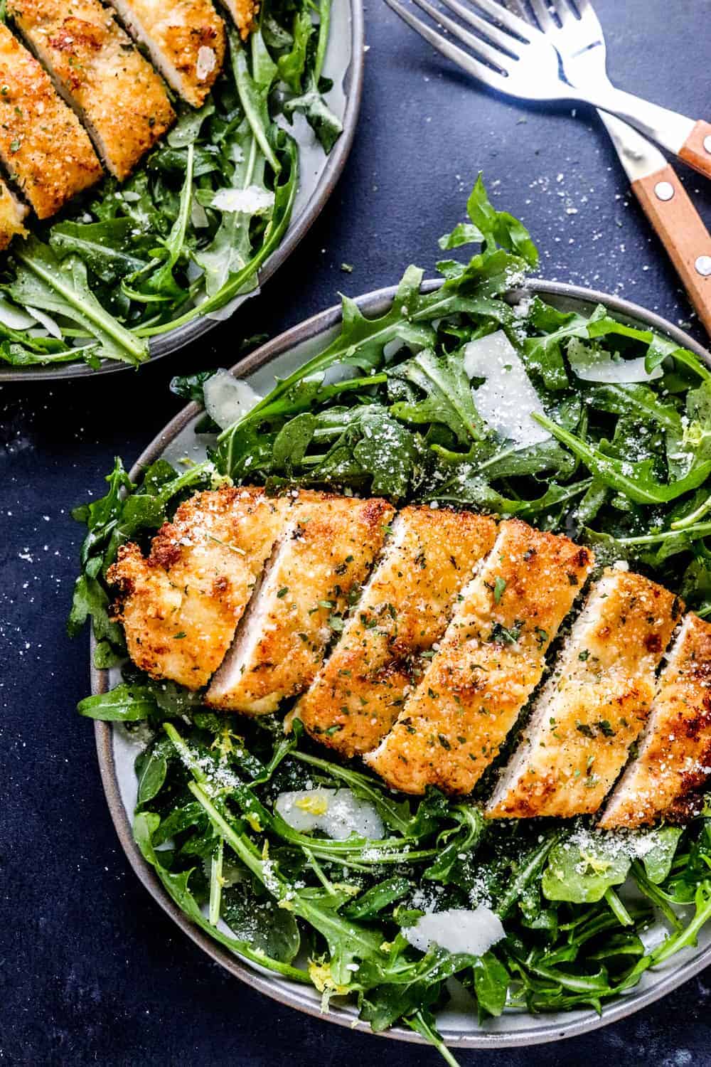 Air fryer breaded chicken breast sliced into strips on a bed of arugula and shaved Parmesan on a plate with another plate of chicken behind it. 